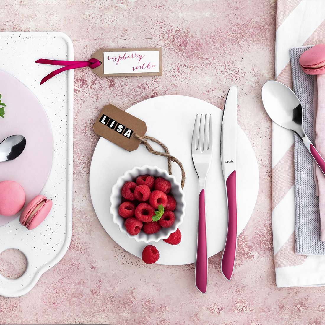 Auf einem rosa gedeckten Tisch sorgen eine Schale mit Himbeeren, buntes Besteck mit rosa Griffen, das Kuppels PRISMA Menümesser Vollheft himbeerrot, Macarons, Namensschilder, eine gestreifte Serviette und ein Etikett mit der Aufschrift "Himbeerwoche" für einen lebendigen Look.