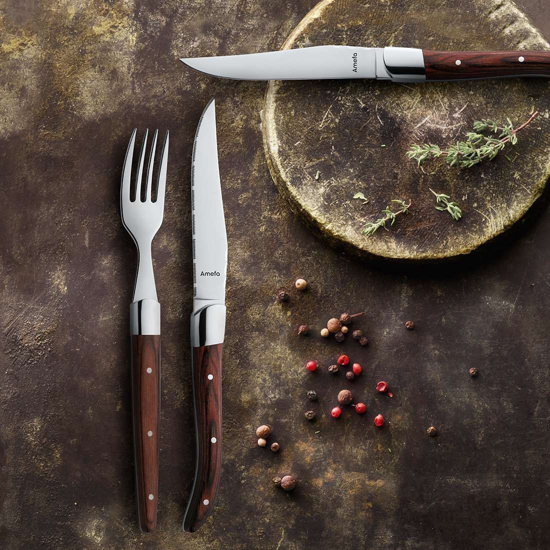 Das Amefa ROYAL STEAK Steakmesser Set 2-teilig holz wird mit einer Gabel auf einer rustikalen braunen Oberfläche neben einem hölzernen Schneidebrett, Kräutern und gemischten Pfefferkörnern präsentiert und vermittelt ein authentisches Steakhouse-Gefühl.