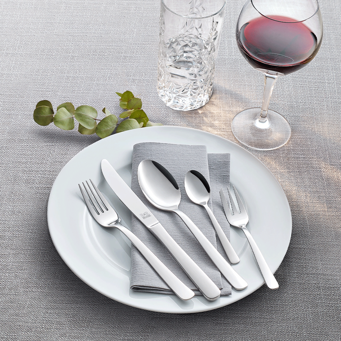 Auf einem weißen Teller mit gefalteter grauer Serviette steht das Beckmann & Rommerskirchen LUNA Besteckset 60-teilig, begleitet von einem Weinglas mit Rotwein, einem Wasserglas aus Kristall und Grünzeug auf einer strukturierten grauen Tischdecke.