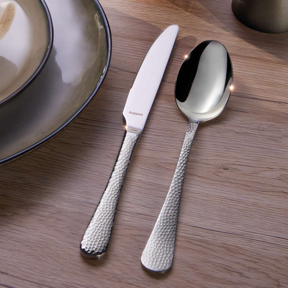 Ein strukturiertes Silbermesser und ein Löffel aus dem Kuppels Home CRYSTAL Besteckset 30-teilig liegen neben Keramiktellern auf einem Holztisch.