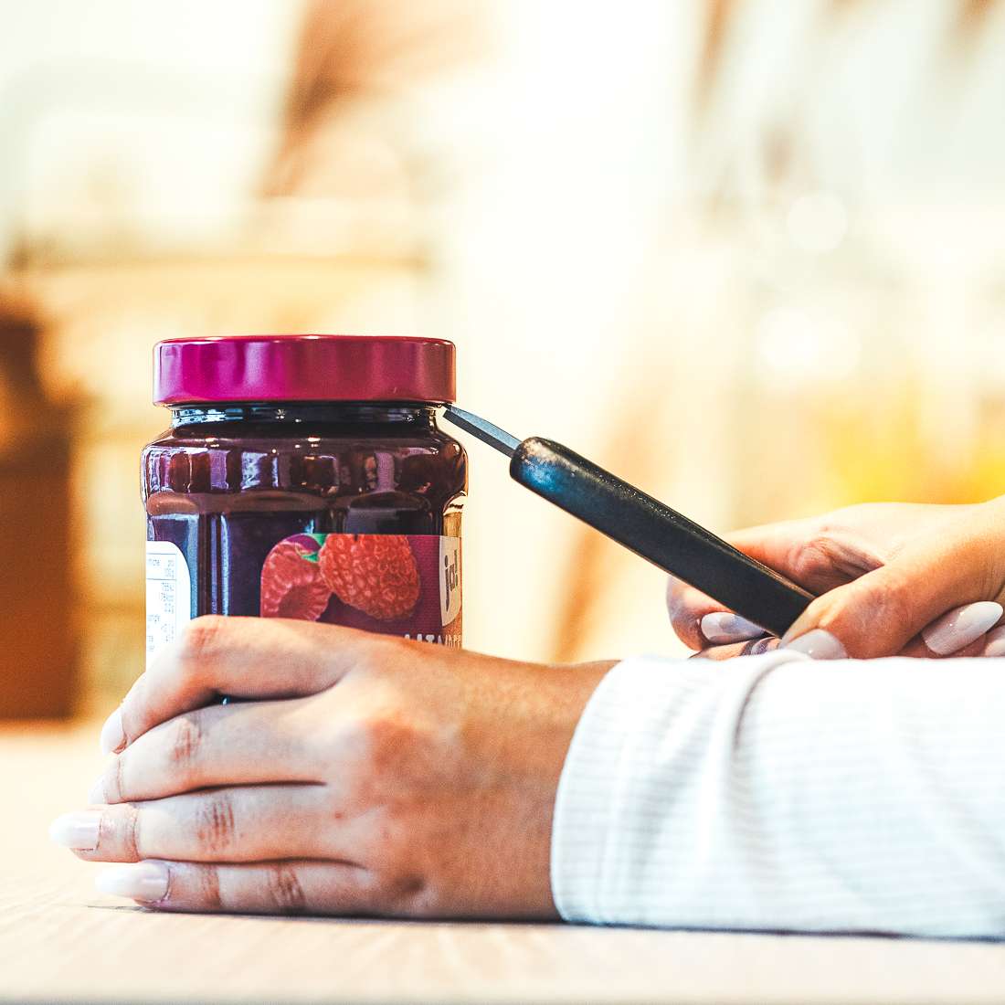 Eine Person benutzt eine Kuppels SELECTION Allzweckschere mit scharfen Klingen, um ein fest verschlossenes Glas Erdbeermarmelade zu öffnen. Dabei hält sie das Glas mit einer Hand fest und hebt den Deckel mit der anderen auf.