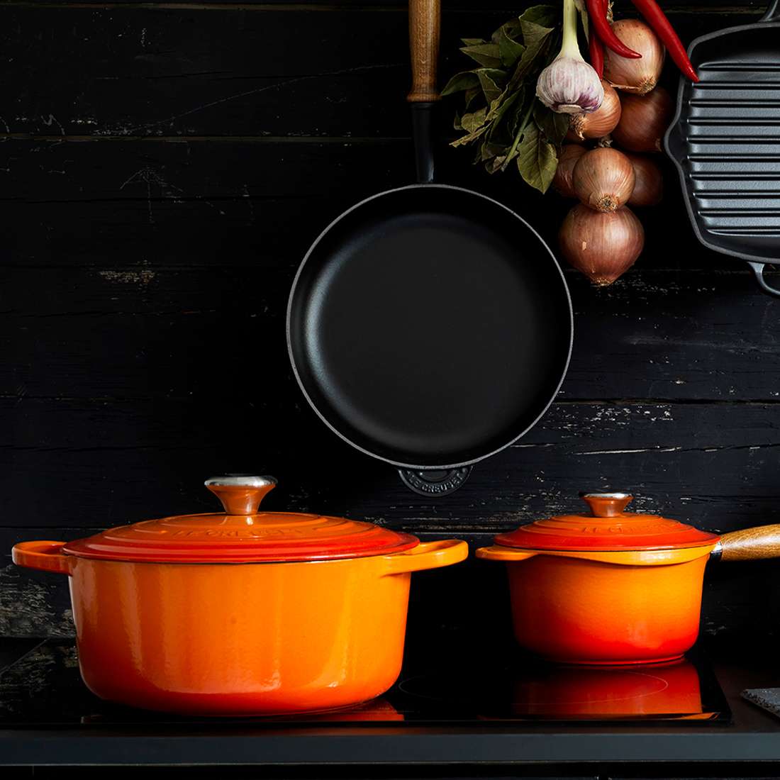 Zwei orangefarbene Le Creuset Signature Bräter rund 28 cm Gusseisentöpfe, einer davon 7 l groß und innen emailliert, stehen auf einer schwarzen Herdplatte. Darüber hängt eine schwarze Bratpfanne mit Zwiebeln, Knoblauch und roten Chilischoten neben einer Grillpfanne auf einem dunklen Holzhintergrund.