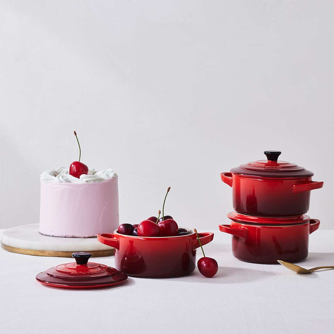 Ein weißer, mit einer Kirsche gekrönter Kuchen steht neben Le Creuset SIGNATURE Mini Cocottes, darunter eine mit Kirschen gefüllte Deckelschale. Ein goldener Löffel liegt nebenan auf einer weißen Tischdecke, bereit zum eleganten Servieren.