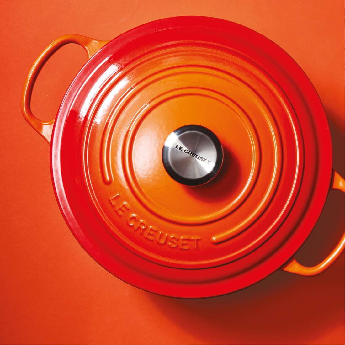 Ein leuchtend orangefarbener Le Creuset Signature Bräter rund 28 cm mit silbernem Knauf sitzt von oben gesehen auf einem orangefarbenen Hintergrund. Der Deckel ist mit der Le Creuset-Prägung versehen und seine emaillierte Innenseite sorgt für perfektes Kochen für bis zu 6 Portionen.