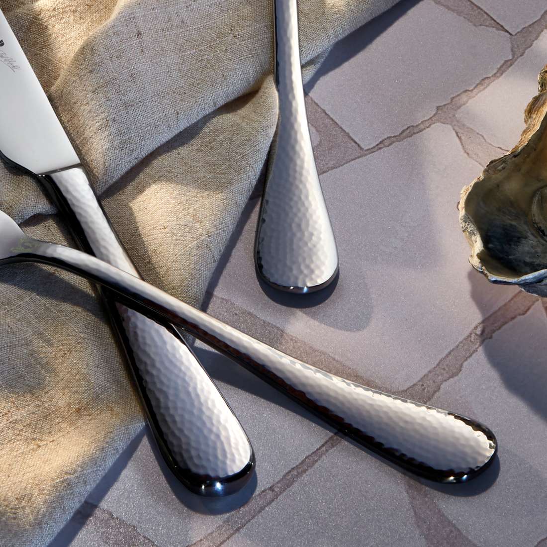 Drei Paul Wirths MONDO Besteckset 30-teilig gehämmerte Metallgriffe im Hammerschlagdesign ruhen auf Steinfliesen, die von einem beigen Stoff und Austernschalen umgeben sind. Sie reflektieren das Licht und heben die strukturierte Edelstahl 18/10 Oberfläche hervor.