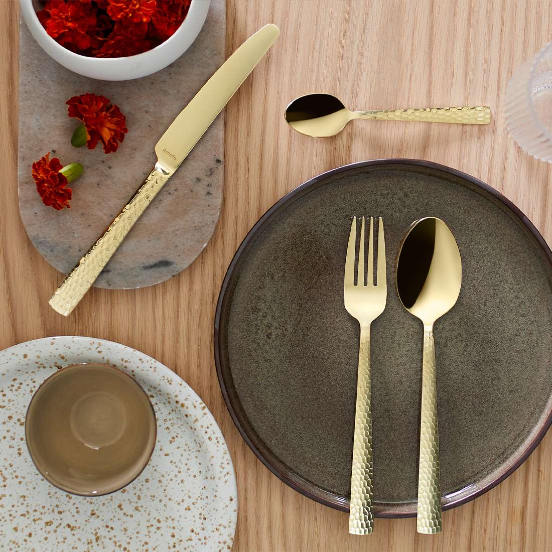 Ein gedeckter Tisch mit dem 6-teiligen Amefa FELICITY PVD Champagner Espressolöffel-Set, einem grauen Teller, einer gesprenkelten Tasse und Untertasse, einem Glas und einem Marmortablett mit roten Blumen auf hellem Holz.