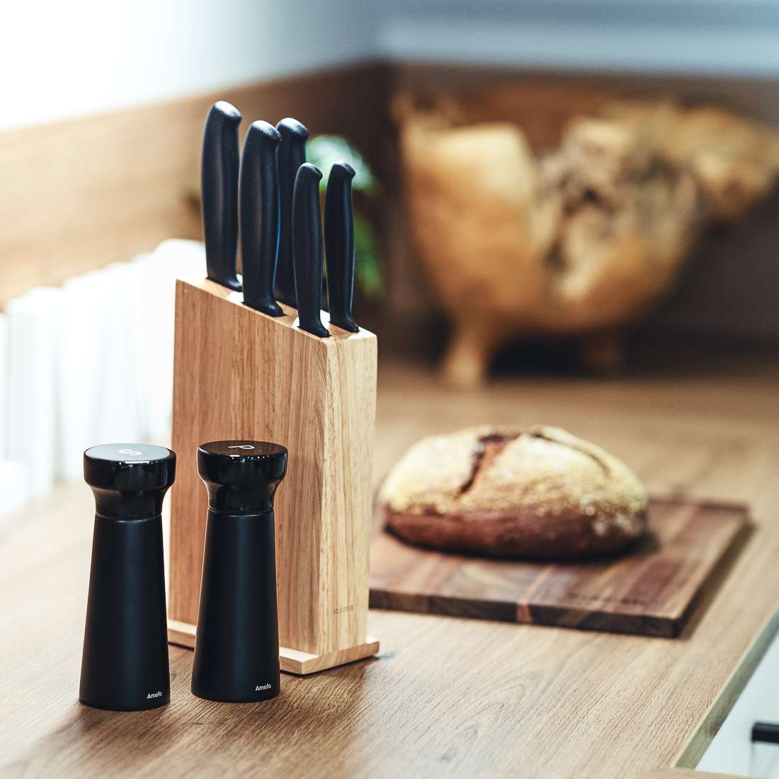 Ein hölzerner Messerblock mit Messern mit schwarzem Griff, das Amefa MODERN WOOD Salz-/Pfeffermühle Set 2-teilig schwarz mit Keramikmahlwerk und ein rustikaler Brotlaib auf einem Schneidebrett stehen auf einer hellbraunen Küchenarbeitsplatte.