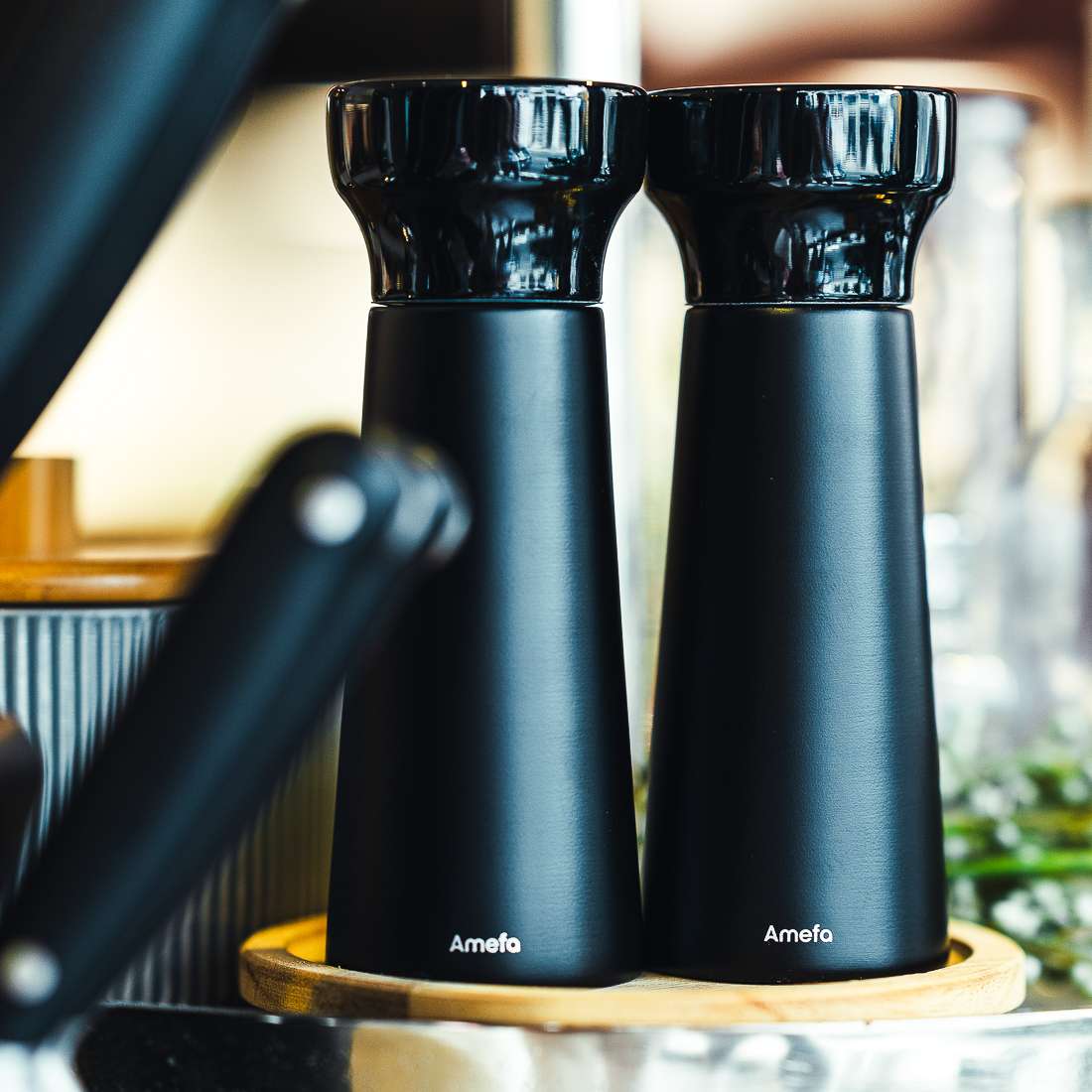Das MODERN WOOD Salz-/Pfeffermühle Set 2-teilig von Amefa umfasst zwei schwarze Mühlen mit glänzenden Köpfen und langlebigem Keramikmahlwerk auf einem runden Holztablett - ideal als Salz- oder Pfeffermühle.