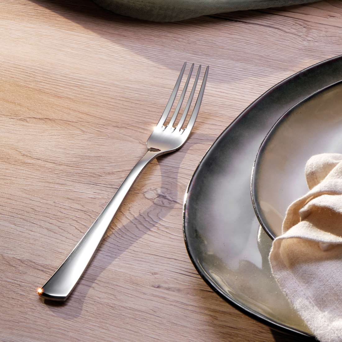 Eine hochglanzpolierte silberne Gabel aus dem Kuppels FLAIR Besteckset 30-teilig liegt auf einem hellen Holztisch neben Keramiktellern und einer gefalteten beigen Serviette. Das Sonnenlicht wirft sanfte Schatten auf die elegante Szene.