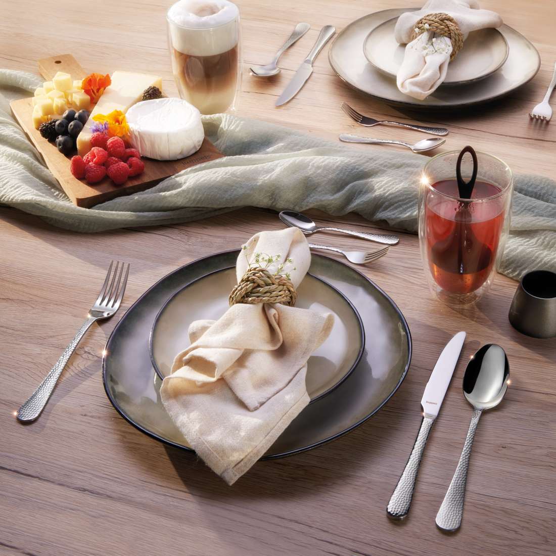 Ein Frühstückstisch für zwei Personen wird mit dem eleganten Kuppels Home CRYSTAL Besteckset 30-teilig, Tellern, Servietten mit Seilring, einer Käse- und Obstplatte, einem Milchkaffee, einem roten Getränk und einem grünen Stoffläufer für einen gemütlichen Holztisch gedeckt.