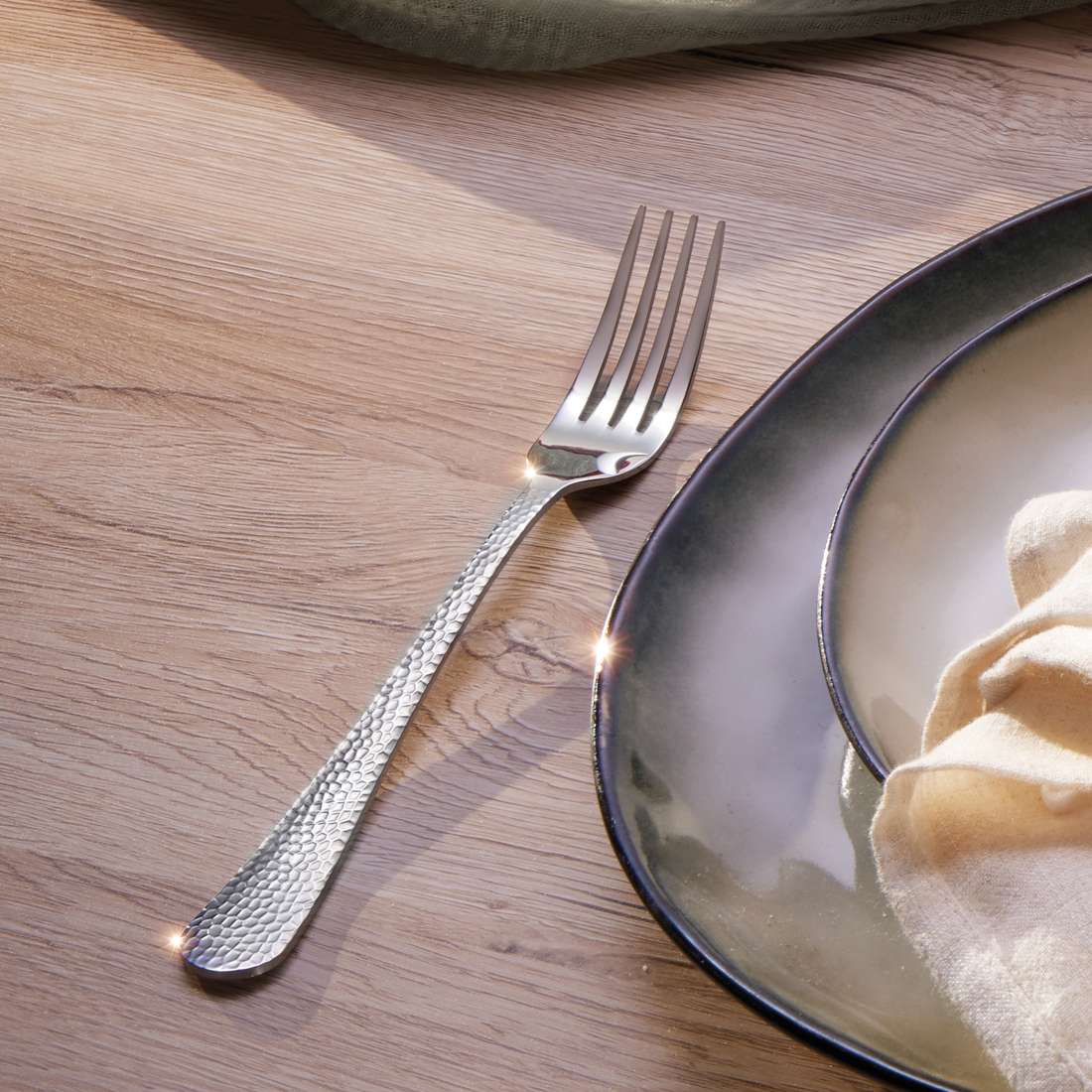 Das CRYSTAL Besteckset 30-teilig von Kuppels Home liegt in Hammerschlag-Optik auf einem Holztisch neben gestapelten grün-grauen Keramiktellern, einer davon mit cremefarbener Serviette, während Sonnenlicht das Besteck zum Glänzen bringt.