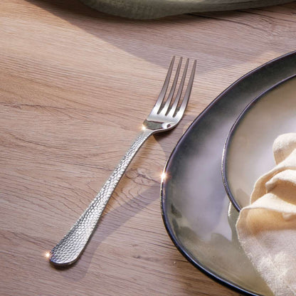 Das CRYSTAL Besteckset 30-teilig von Kuppels Home liegt in Hammerschlag-Optik auf einem Holztisch neben gestapelten grün-grauen Keramiktellern, einer davon mit cremefarbener Serviette, während Sonnenlicht das Besteck zum Glänzen bringt.