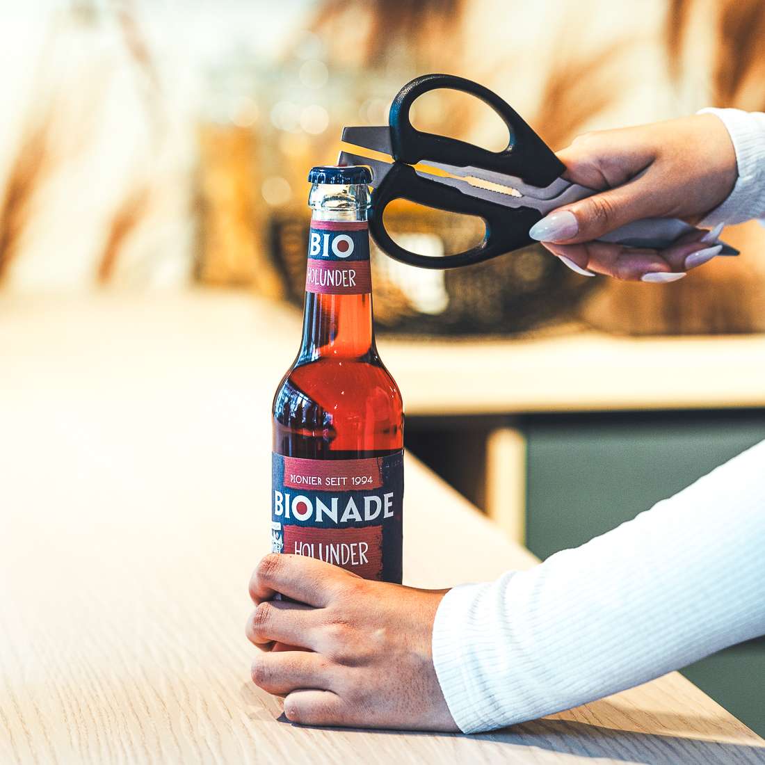 Eine Person öffnet mit einer Kuppels SELECTION Allzweckschere eine Flasche Bionade Holunder Getränk auf einem hellen Holztisch und trägt ein weißes langärmeliges Oberteil.