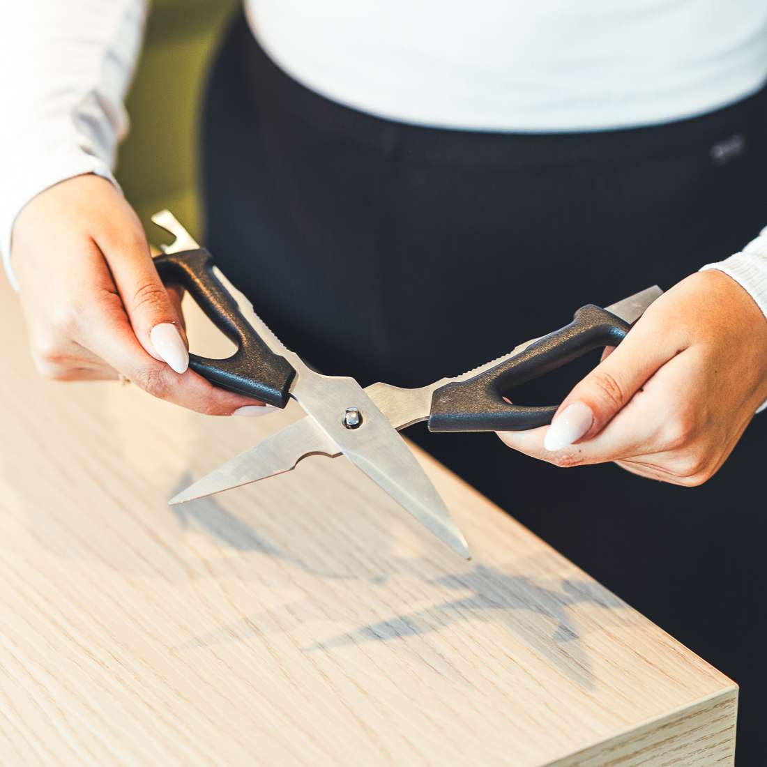 Eine Person mit hellem Nagellack hält eine Kuppels SELECTION Allzweckschere mit schwarzen Griffen und scharfen Klingen über eine helle Holzoberfläche.