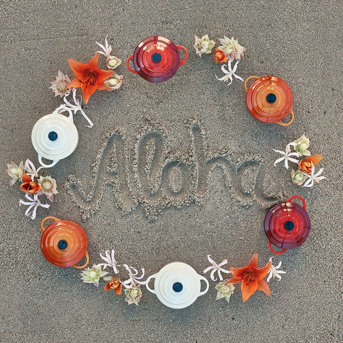 Das Wort "Aloha" ist in Sand geschrieben, umgeben von orangefarbenen Blumen, weißen Seesternen und der kreisförmig angeordneten Le Creuset SIGNATURE Mini Cocotte - perfekt zum Servieren Ihrer Lieblingsspeisen.