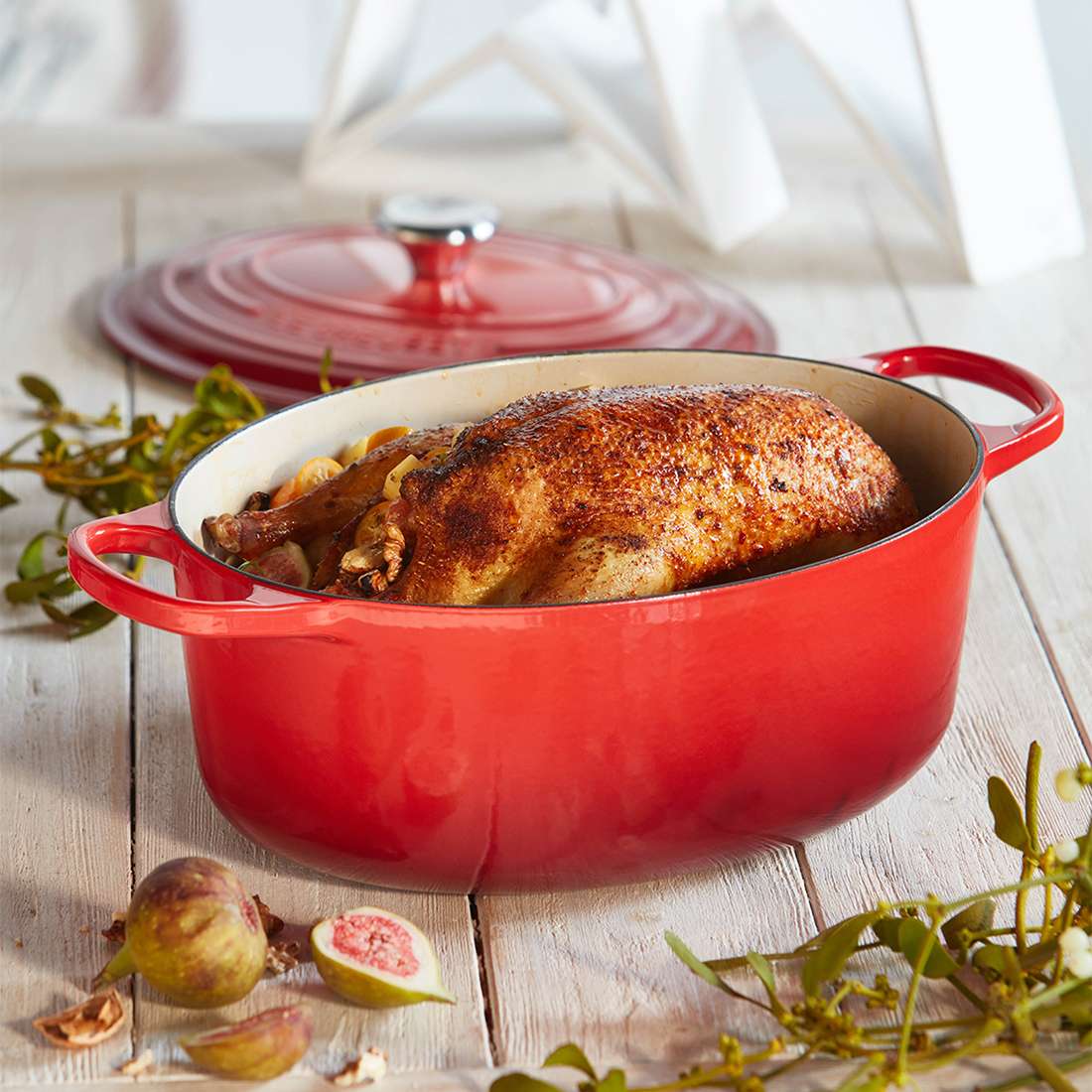 Ein ganzes Brathähnchen ruht in einem Le Creuset Signature Bräter oval 29 cm mit emaillierter Innenseite auf einem weißen Holztisch, umgeben von Feigen und grünen Blättern, mit dem dichtschließenden Deckel daneben.