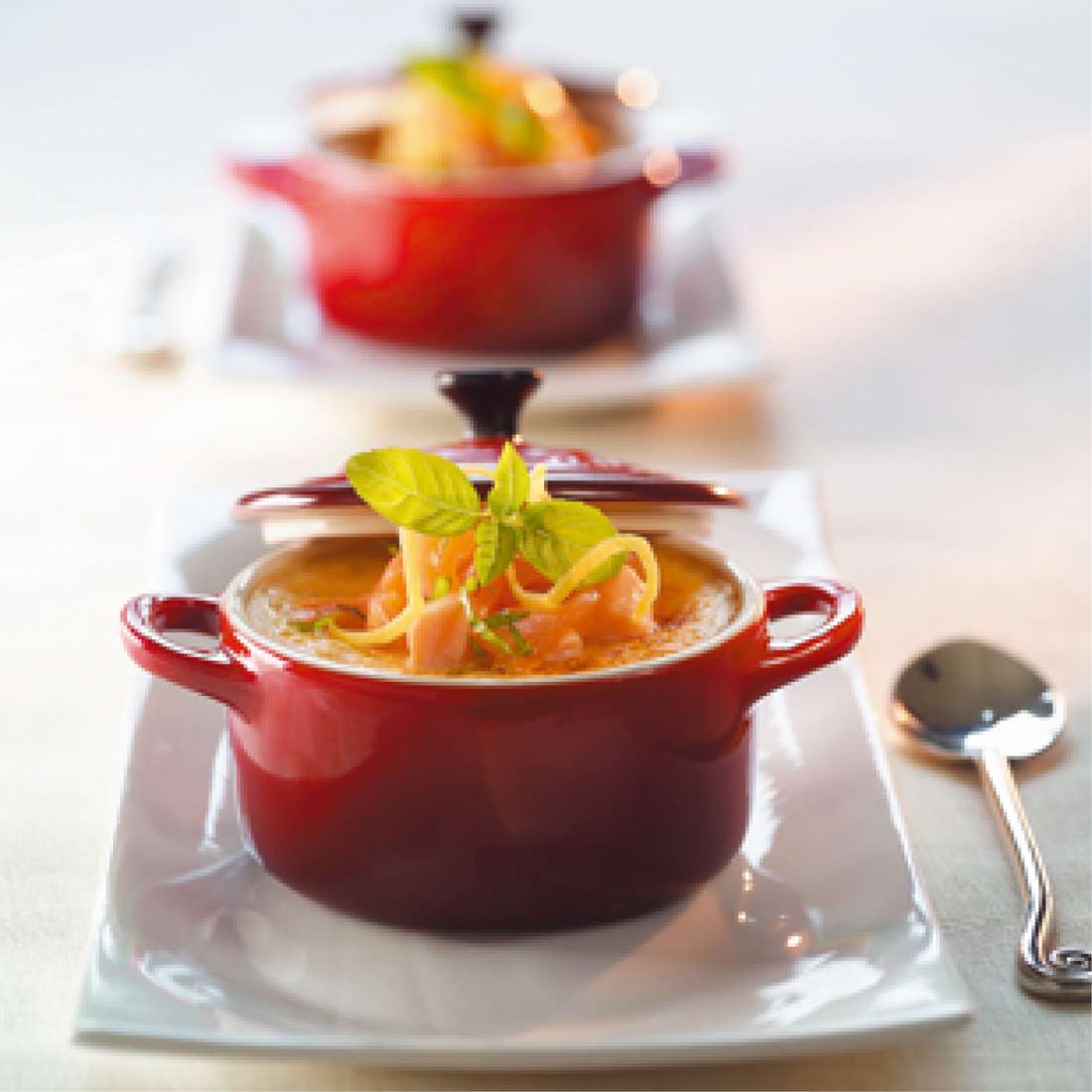 Eine rote Le Creuset SIGNATURE Mini Cocotte, gefüllt mit Suppe, garniert mit Kräutern und Gemüse, steht auf einem weißen rechteckigen Teller mit einem Löffel daneben; eine weitere Mini Cocotte ist im Hintergrund verschwommen.