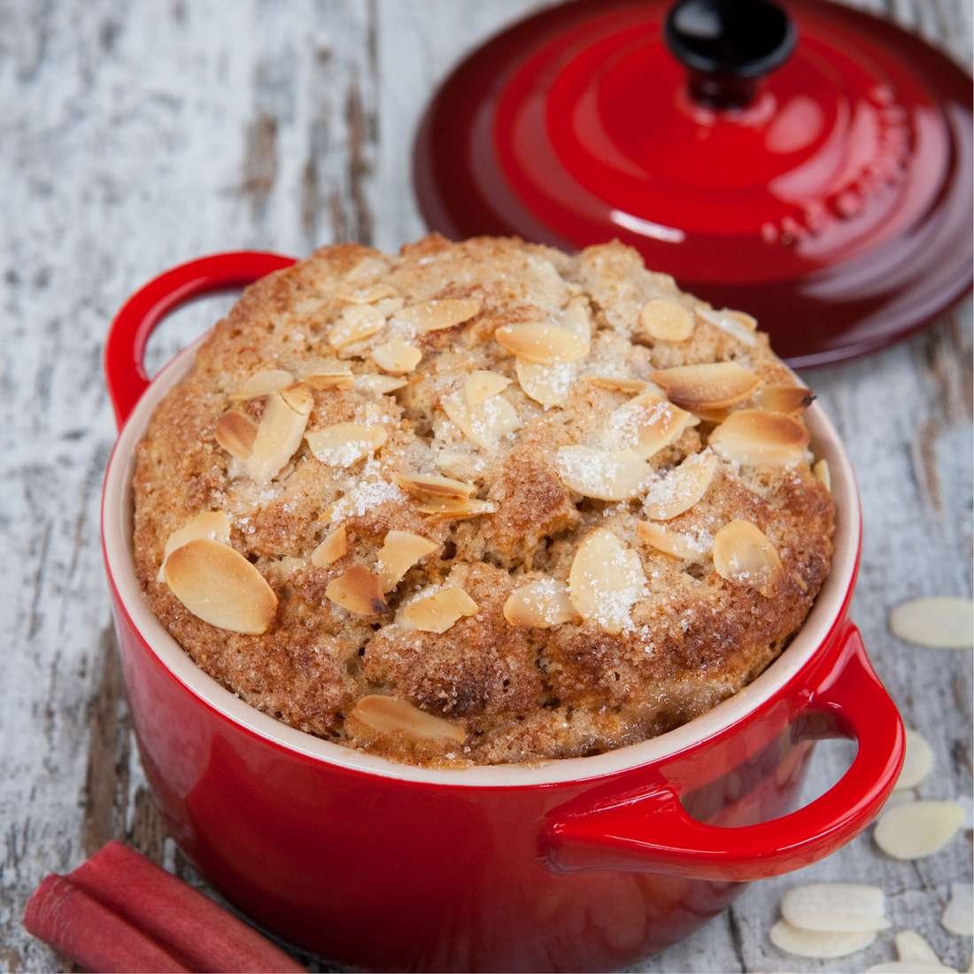 Ein goldbrauner Mandelkuchen mit gehobelten Mandeln liegt in einer Le Creuset SIGNATURE Mini Cocotte mit Griffen servierfertig neben dem passenden roten Deckel auf einer rustikalen Holzfläche.