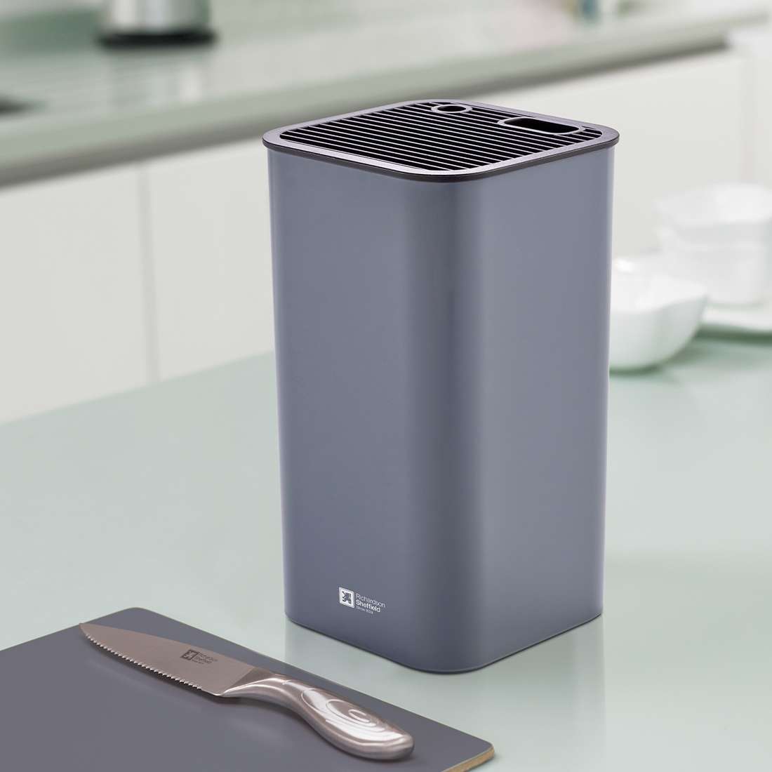 Der rechteckige, graue Messerblock universal von Richardson Sheffield steht platzsparend auf der hellen Arbeitsplatte neben einem Schneidebrett, im Hintergrund sieht man eine unscharfe Fläche und weiße Schüsseln.