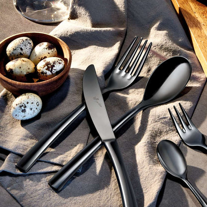 Das SWING Besteckset 4-teilig PVD schwarz von Paul Wirths-Gabel, Messer, Löffel und Teelöffel-liegt auf beigem Tuch neben einer Holzschale mit Wachteleiern, daneben sind ein Glas und ein Holzbrett zu sehen.